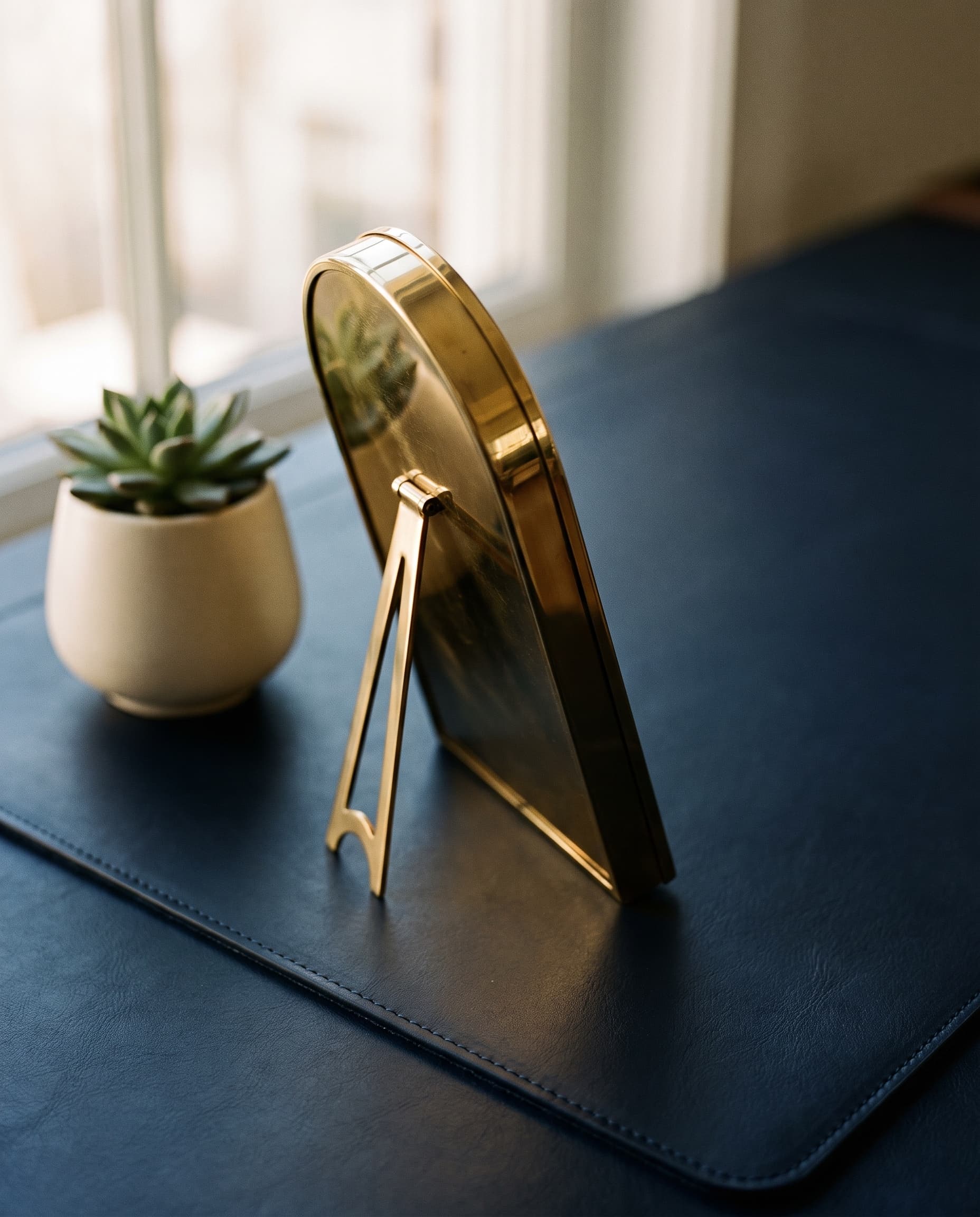 An arched brass frame on its easel stand and a small succulent in an ivory pot, on a navy leather desk pad by a sunny window