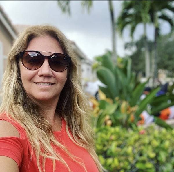 Maria Iraola in sunglasses and a coral top, smiling outdoors among tropical plants