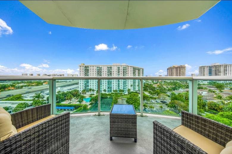 Balcony with wicker armchairs and a glass railing, overlooking condo towers and palm trees